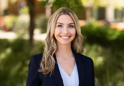 Professional outdoor headshot of a woman with long blonde hair wearing a dark blazer over a light-colored blouse. She is smiling at the camera while standing in a landscaped setting with trees and greenery in the softly blurred background. Natural lighting and a shallow depth of field create a polished, professional portrait.