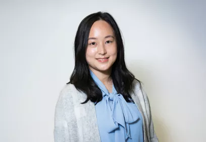 Professional portrait of a woman with shoulder-length dark hair wearing a light blue blouse with a bow tie neckline and a light gray cardigan. She is smiling gently at the camera while standing against a plain light gray background.