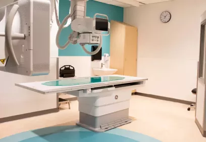 Modern X-ray imaging room featuring a digital radiography table and overhead X-ray equipment. The clean clinical space includes a sink, wall clock, and patient positioning area, highlighting a contemporary medical imaging environment.