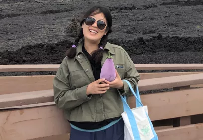 Xiaoxi Liu smiles while standing on a viewing platform in front of a volcanic lava field. Wearing sunglasses and a green jacket, she holds a small purple plush toy and carries a tote bag, with the rugged landscape visible behind her.
