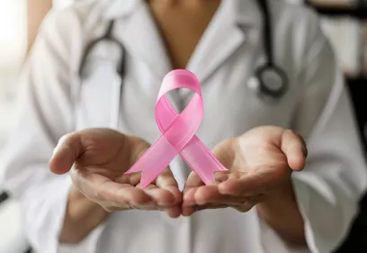 A healthcare professional in a white coat holds a pink breast cancer awareness ribbon in outstretched hands. A stethoscope hangs around their neck, and the softly blurred background keeps the focus on the ribbon, symbolizing support, awareness, and advocacy for breast cancer patients and survivors.