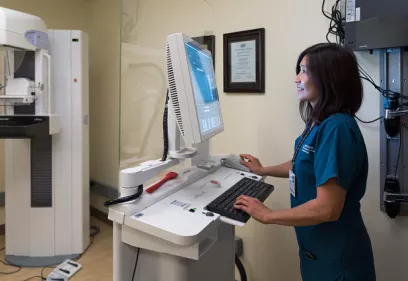 Healthcare professional operating a mammography imaging workstation in a breast imaging clinic. The technologist is seated at a computer console, viewing images on a monitor while using a keyboard and mouse. A mammography machine is visible in the examination room behind a glass partition. The clinical setting is clean and modern, highlighting the technology and expertise involved in breast cancer screening and diagnostic imaging.