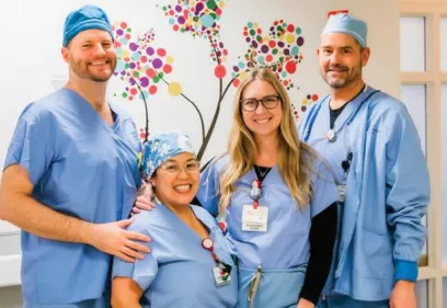 Group photo of four healthcare professionals standing together in a hospital or clinical setting. The team is dressed in blue medical scrubs, with two members also wearing surgical caps. They are smiling toward the camera and standing in front of a colorful wall mural depicting a tree made of multicolored circular leaves. Identification badges are visible on several team members, and one person has a hand resting on a colleague’s shoulder. The image conveys teamwork, compassion, and a supportive healthcare.