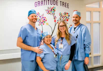 Four healthcare professionals in surgical scrubs stand together smiling in front of a colorful "Gratitude Tree" wall display. The group poses in a hospital hallway, highlighting teamwork, appreciation, and a positive workplace culture.