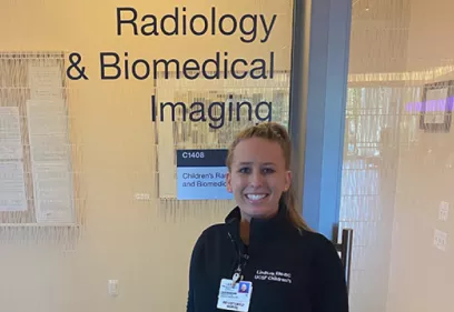 A healthcare professional stands smiling in front of a glass entrance displaying the words "Radiology & Biomedical Imaging." Wearing a UCSF Children's identification badge and black jacket, she poses in a clinical facility, highlighting her role within the radiology department.
