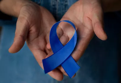 Two open hands gently holding a blue awareness ribbon. The ribbon rests in the center of the palms, symbolizing support and awareness. The background is softly blurred, drawing attention to the ribbon and hands. The blue ribbon is commonly associated with prostate cancer awareness.