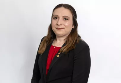 Professional portrait of a woman wearing a black blazer over a red blouse. She is standing against a light gray background and looking at the camera with a slight smile. Small lapel pins are visible on her blazer, giving the image a polished and professional appearance.