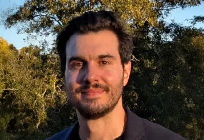 A head-and-shoulders portrait of a person standing outdoors in warm evening sunlight. He has dark hair, a short beard, and is wearing a dark jacket over a black shirt. Trees and foliage fill the softly blurred background, creating a natural setting.