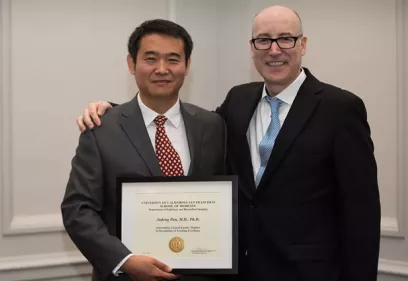 Two people in business attire stand side by side, smiling at the camera. The person on the left holds a framed award certificate while the person on the right has an arm around their shoulder in congratulations. Both are wearing suits and ties, and the photo is taken indoors against a neutral background, marking an award or recognition ceremony.