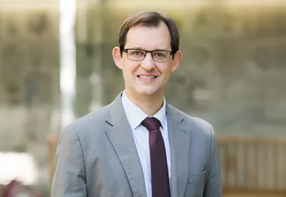 A professionally dressed person wearing a light gray suit, white shirt, and dark burgundy tie smiles at the camera. They have short dark hair and rectangular glasses. The portrait is taken outdoors or in a bright atrium, with a softly blurred background featuring glass panels and a wooden bench.