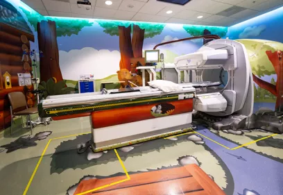A hospital MRI suite decorated with a colorful forest-themed mural featuring trees, wildlife, and mountains. The MRI scanner and patient table are surrounded by child-friendly artwork designed to create a calming, welcoming environment for pediatric patients. Medical equipment, a monitoring station, and seating are visible within the brightly lit room.