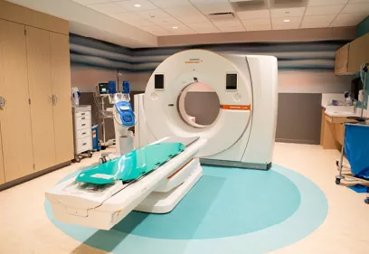 A modern CT imaging suite featuring a large white CT scanner with an attached patient table positioned in the center of the room. Medical equipment, cabinets, and a patient monitoring station surround the scanner, while the clean, brightly lit space has wood cabinetry, teal flooring accents, and a calming striped wall design.