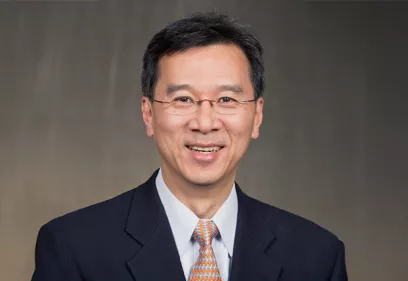 Professional headshot of a smiling man wearing glasses, a dark suit, a light blue dress shirt, and an orange patterned tie, posed against a neutral gray background.