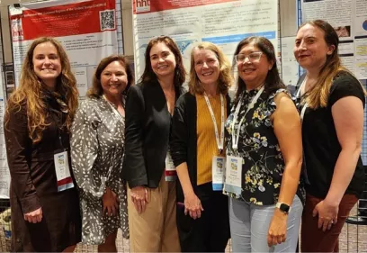 Six conference attendees stand together smiling in front of research posters at a scientific poster session. All are wearing conference badges, and the posters are displayed on boards in the background.
