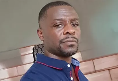 A head-and-shoulders portrait of a person wearing a navy blue polo shirt with red trim. He is looking toward the camera with a neutral expression while standing indoors, with a tiled wall and softly blurred background behind him.