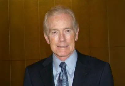 A professional head-and-shoulders portrait of a person wearing a dark suit, light blue dress shirt, and patterned blue tie. He has short gray hair and a slight smile and is facing the camera. The background is a warm-toned wood-paneled wall, giving the image a formal, indoor setting.
