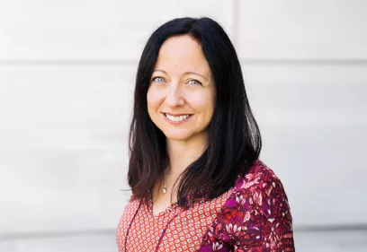 Janine Lupo smiles at the camera in a professional headshot, wearing a patterned red and pink blouse against a light stone background.