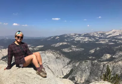 A woman wearing sunglasses, hiking boots, and outdoor clothing sits on a rocky overlook with a sweeping view of granite cliffs, forested mountains, and a clear blue sky in the background.