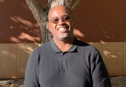 Jerome Taylor smiles outdoors wearing a black polo shirt and sunglasses, standing in front of a softly blurred background of trees and greenery.