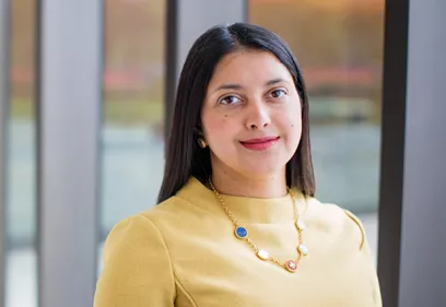 A professional head-and-shoulders portrait of a person standing indoors in front of large floor-to-ceiling windows. She is wearing a light yellow long-sleeve top, small gold earrings, and a gold necklace with blue, white, and red circular accents. She faces the camera with a slight smile. The softly blurred background shows glass panels and an outdoor landscape, creating a bright, modern office or campus setting.
