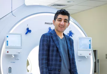 Professional portrait of a smiling man wearing a blue plaid blazer over a blue shirt, standing in front of an MRI scanner in a medical imaging suite. The scanner and its control displays are visible in the background.