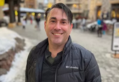 Professional portrait of a smiling man wearing a black jacket over a dark shirt, standing on a snowy pedestrian street with shops and buildings softly blurred in the background.