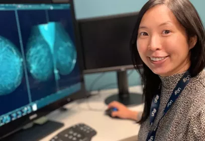 A smiling healthcare professional sits at a computer workstation, looking toward the camera while using a mouse. She is wearing a patterned long-sleeve top and a UCSF-branded lanyard. Two large monitors display grayscale breast mammography images. The photo appears to be taken in a clinical imaging or radiology reading room with additional monitors and office equipment visible in the background.