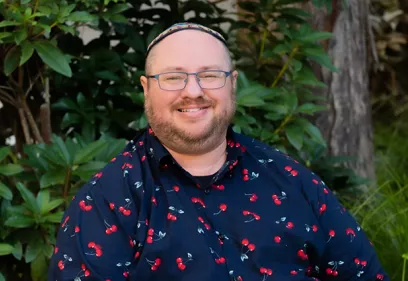 A professional head-and-shoulders portrait of a smiling person seated outdoors in front of lush green foliage. He is wearing glasses, a dark button-up shirt with a red cherry pattern, and a colorful skullcap. He is facing the camera with a friendly expression in a natural, softly blurred setting.