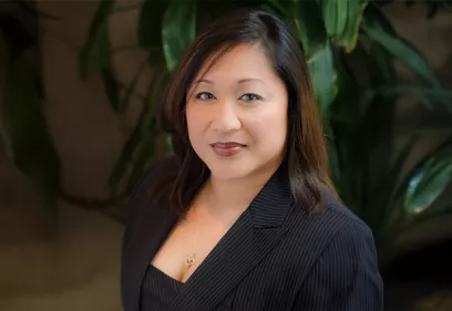 A professional head-and-shoulders portrait of a person wearing a dark pinstripe blazer and matching top. She is facing the camera with a slight smile, wearing a delicate necklace, and standing in front of softly blurred green foliage in an indoor setting.