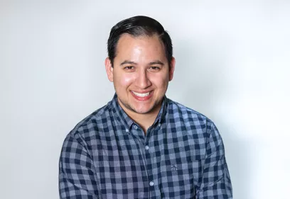 Professional headshot of a smiling man wearing a blue and gray checkered button-down shirt against a light gray background.