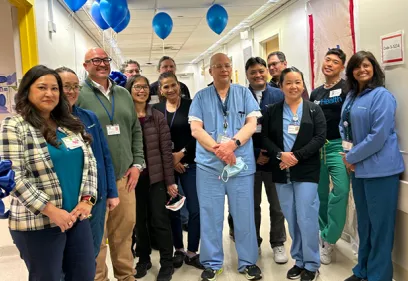 A group of healthcare staff pose together in a hospital hallway during a ribbon-cutting celebration. Blue balloons and smiles mark the occasion, with clinicians and staff wearing a mix of scrubs, lab coats, and business attire.