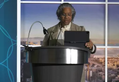 A presenter stands behind a black podium, speaking while looking at a tablet mounted on the lectern. She is wearing a light-colored blouse with a blazer and has gray hair and glasses. A microphone and reading lamp are attached to the podium. Behind her is a large backdrop featuring a city skyline and waterfront, suggesting a conference or auditorium setting.