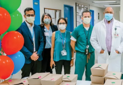 Five masked healthcare professionals stand together in a hospital hallway behind a table with boxed meals. Colorful balloons decorate the scene, suggesting a celebration or recognition event.