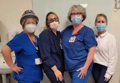 Four healthcare professionals stand together in a clinical room, smiling toward the camera. They are wearing scrubs or clinical attire, face masks, and ID badges, with one person also wearing a decorated surgical cap. A sink and medical equipment are visible in the background, indicating a hospital or patient care setting.