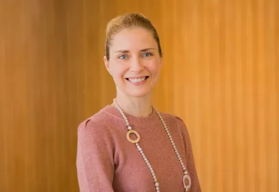 A professional head-and-shoulders portrait of a smiling person standing in front of a warm-toned wood-paneled background. She is wearing a rose-colored long-sleeve top and a long beaded necklace with circular accents. Her hair is pulled back, and she is facing the camera with a friendly expression.