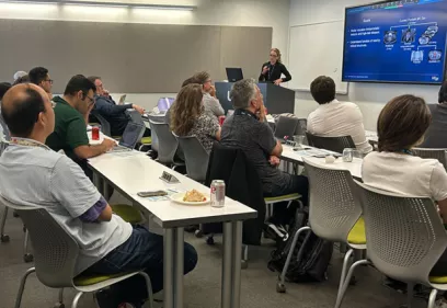 A presenter stands at the front of a conference room delivering a presentation to an audience seated at tables. Attendees listen while viewing a large screen displaying slides about prostate cancer imaging and artificial intelligence. Laptops, notebooks, and refreshments are visible, highlighting an educational symposium or professional training session.