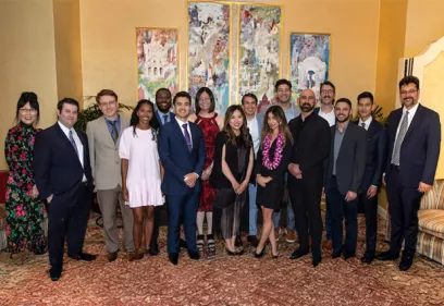 A group of 15 people pose together for a formal photograph in an elegant room with gold walls, patterned carpet, and large framed paintings in the background. The group is dressed in business and semi-formal attire, with several people wearing suits and dresses, and they are smiling while facing the camera.