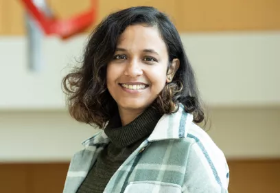 Rupsa Bhattacharjee smiling in an indoor portrait, wearing a green turtleneck and a light plaid overshirt.