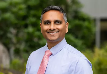 Professional headshot of a smiling man wearing a light blue dress shirt and a pink patterned tie, standing outdoors with blurred green foliage in the background.
