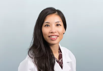 Shirley Chou, MD, wearing a white lab coat, smiles at the camera in a professional portrait against a light blue background.