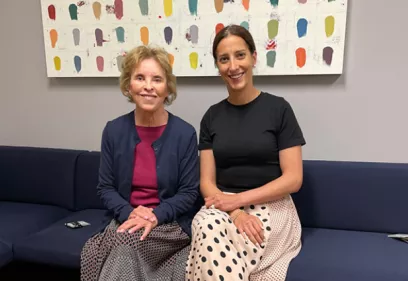 Two women sit together on a blue bench beneath a colorful abstract painting, smiling at the camera.