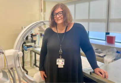 Professional portrait of a woman wearing glasses, a black dress, and a hospital ID badge, standing in an interventional radiology procedure room beside medical imaging equipment. Hospital equipment, a patient bed, and large windows are visible in the background.