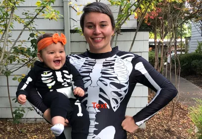 A smiling adult holds a laughing young child outdoors in front of a house and trees. Both are wearing matching black skeleton-themed Halloween costumes, with the child also wearing a bright orange headband. The adult has one hand on their hip while the child smiles toward the camera.