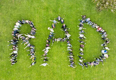 Aerial view of dozens of people lying on a grassy lawn to spell "RAD," with a few people standing in the center and flowering trees visible at the edge of the image.