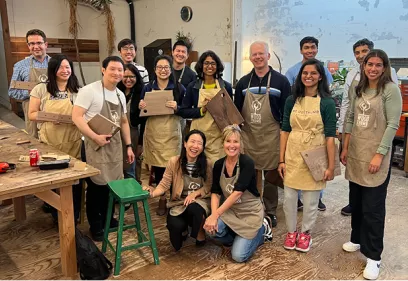 A group of 12 people wearing tan woodworking aprons pose together inside a woodworking studio, smiling and holding handmade wooden cutting boards. Two people kneel in the front while the others stand behind them. Workbenches, tools, unfinished wood projects, and workshop equipment are visible in the background, highlighting a collaborative team-building woodworking event.