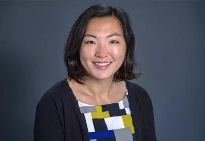 A professional head-and-shoulders portrait of a smiling person against a solid dark gray background. She has shoulder-length dark hair and is wearing a black cardigan over a geometric-patterned blouse in white, gray, black, yellow, and blue. She faces the camera with a friendly expression in a studio-style photograph.