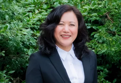 A professional head-and-shoulders portrait of a smiling person standing outdoors in front of lush green foliage. She is wearing a black blazer over a white collared shirt and faces the camera with a friendly expression. The softly blurred greenery provides a natural background.