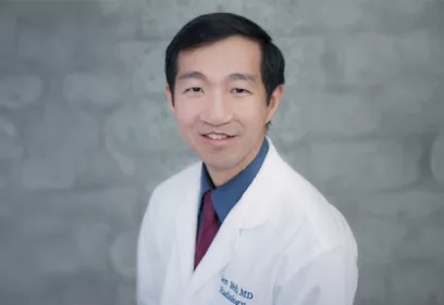 A professional head-and-shoulders portrait of a smiling physician wearing a white medical coat over a blue shirt and burgundy tie. He is facing the camera against a softly blurred gray background, creating a clean, studio-style portrait.