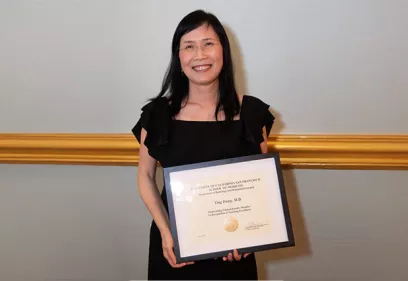 A woman in a black dress smiles while holding a framed certificate of recognition. She stands against a gray wall with gold trim, posing for an award presentation photo.