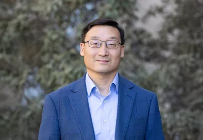 Portrait of a man wearing glasses, a blue blazer, and a light blue dress shirt, standing outdoors in front of softly blurred evergreen trees and looking at the camera with a slight smile.